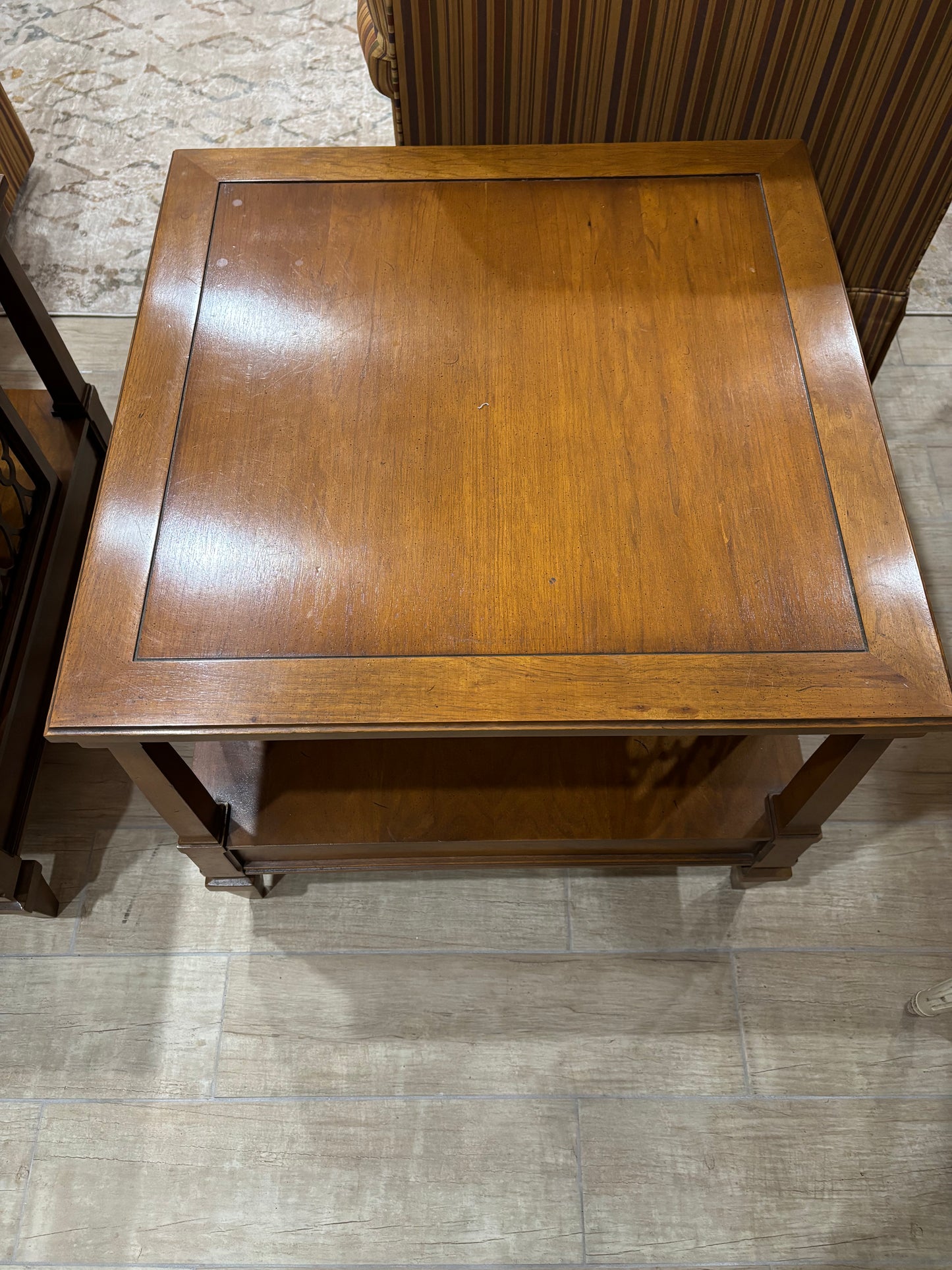 A square wooden end table with a warm medium-brown finish, featuring a recessed panel on top and a lower shelf, photographed from above on a light wood floor.