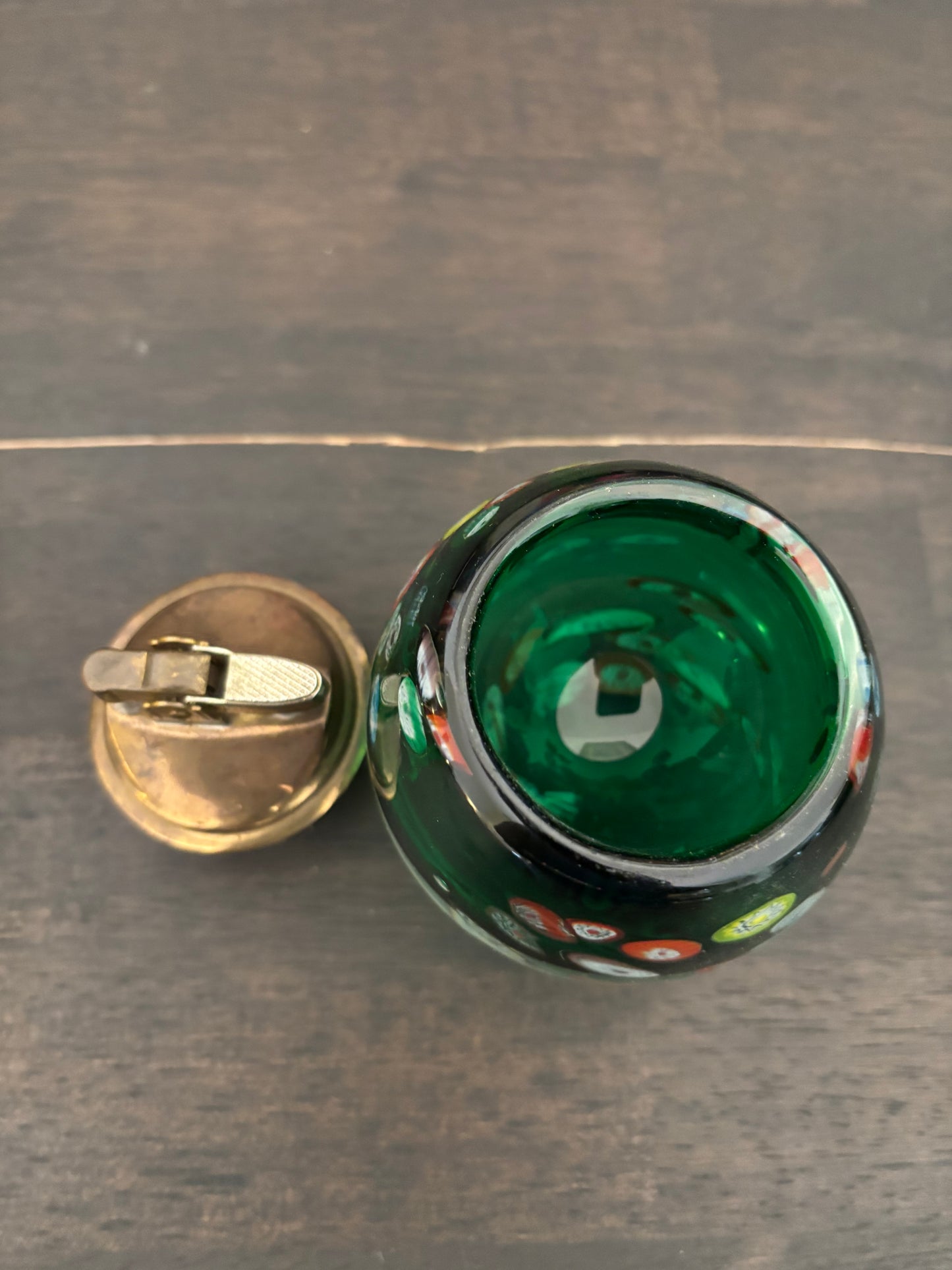 A close-up top-down view of a green glass container or candle holder with a separate brass-toned metal lid resting beside it on a dark wood surface.
