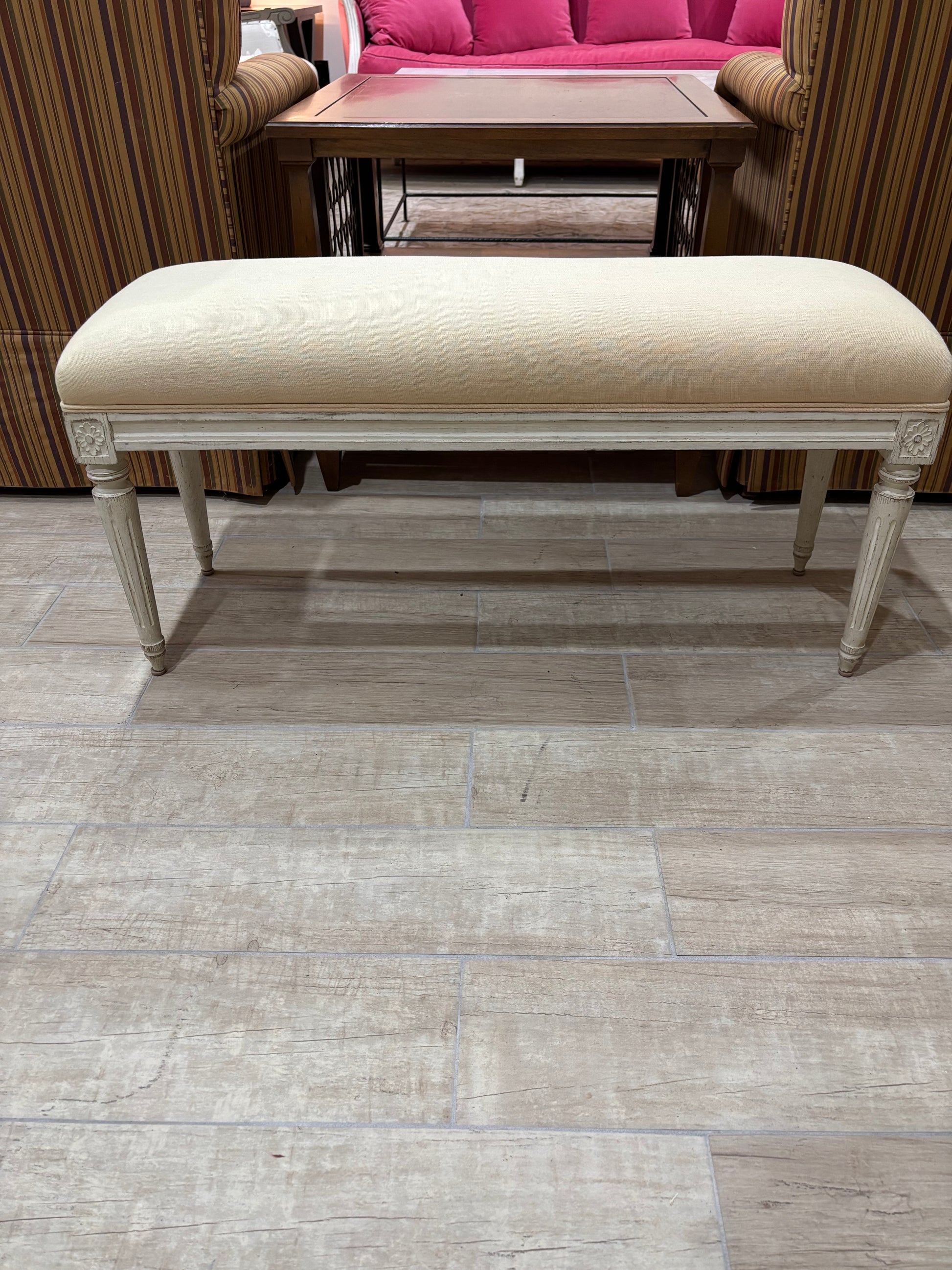 A cream upholstered bench with a distressed off-white frame and turned legs, positioned on a light gray wood-look tile floor in a showroom setting.