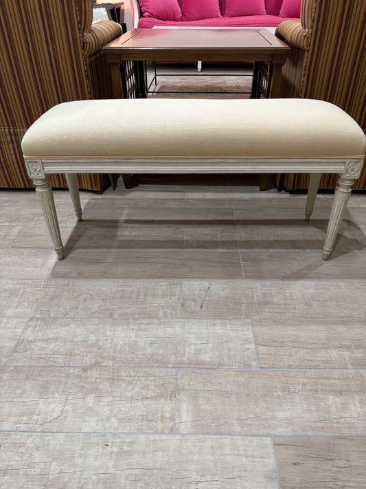 A cream upholstered bench with a distressed off-white frame and turned legs, positioned on a light gray wood-look tile floor in a showroom setting.