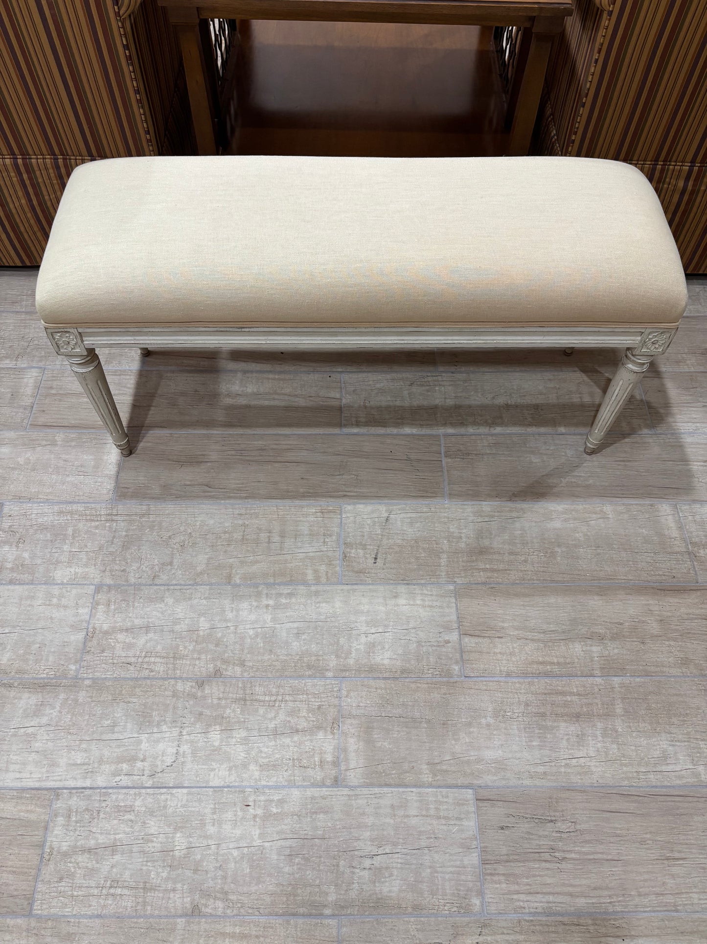 An upholstered bench with a cream fabric cushion and a distressed light gray frame, positioned on a gray wood-look tile floor in front of a dark wood cabinet.