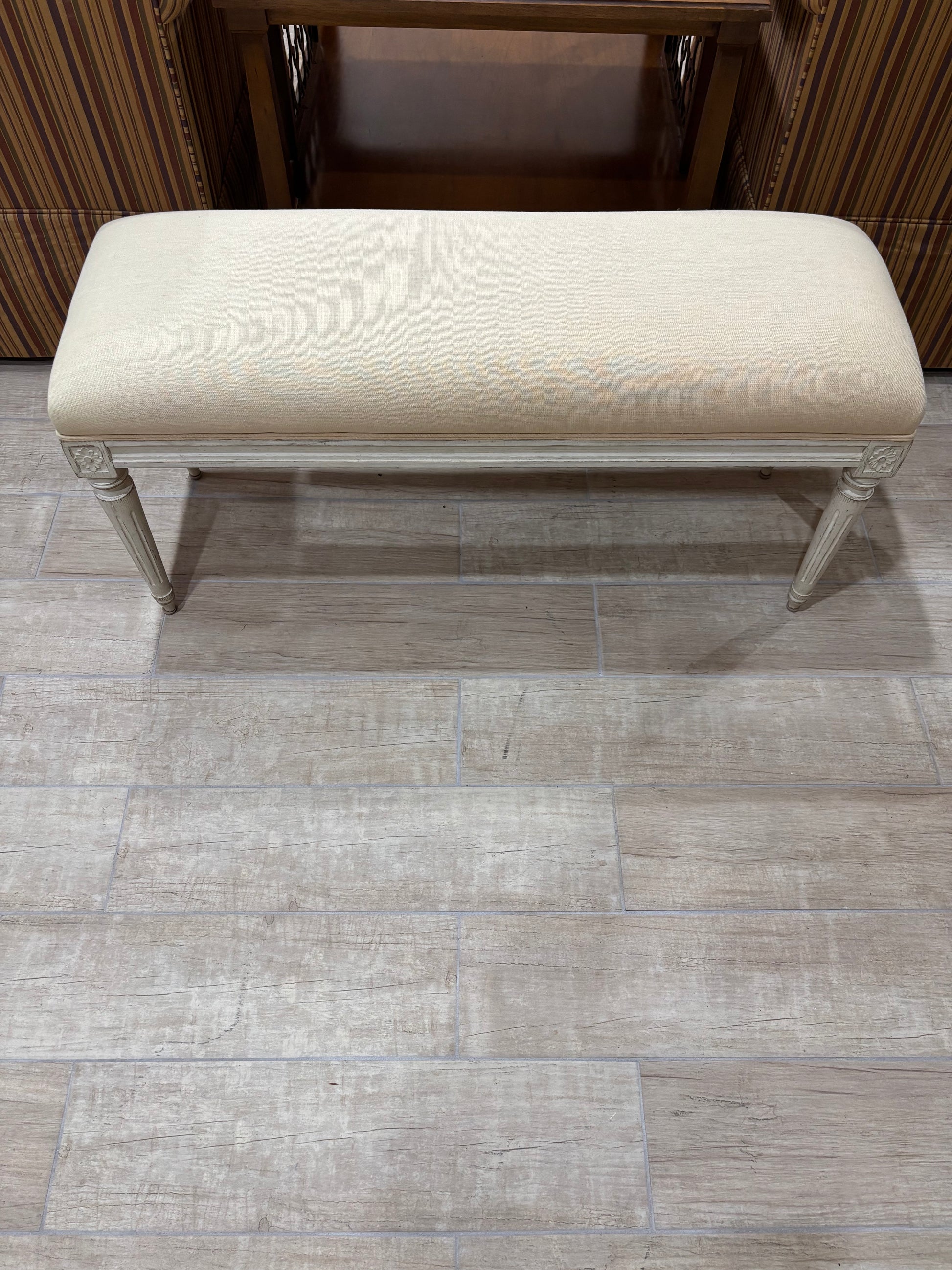 An upholstered bench with a cream fabric cushion and a distressed light gray frame, positioned on a gray wood-look tile floor in front of a dark wood cabinet.