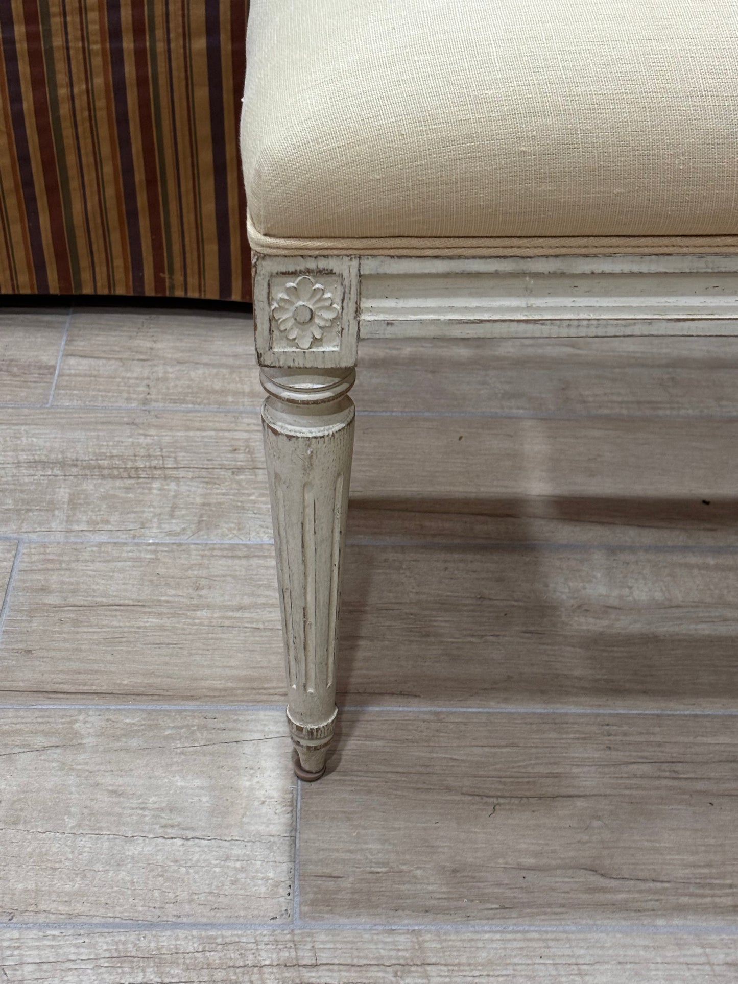 Close-up of a bench or stool leg with a distressed off-white painted frame and a beige upholstered seat, shown on a light wood-look floor.