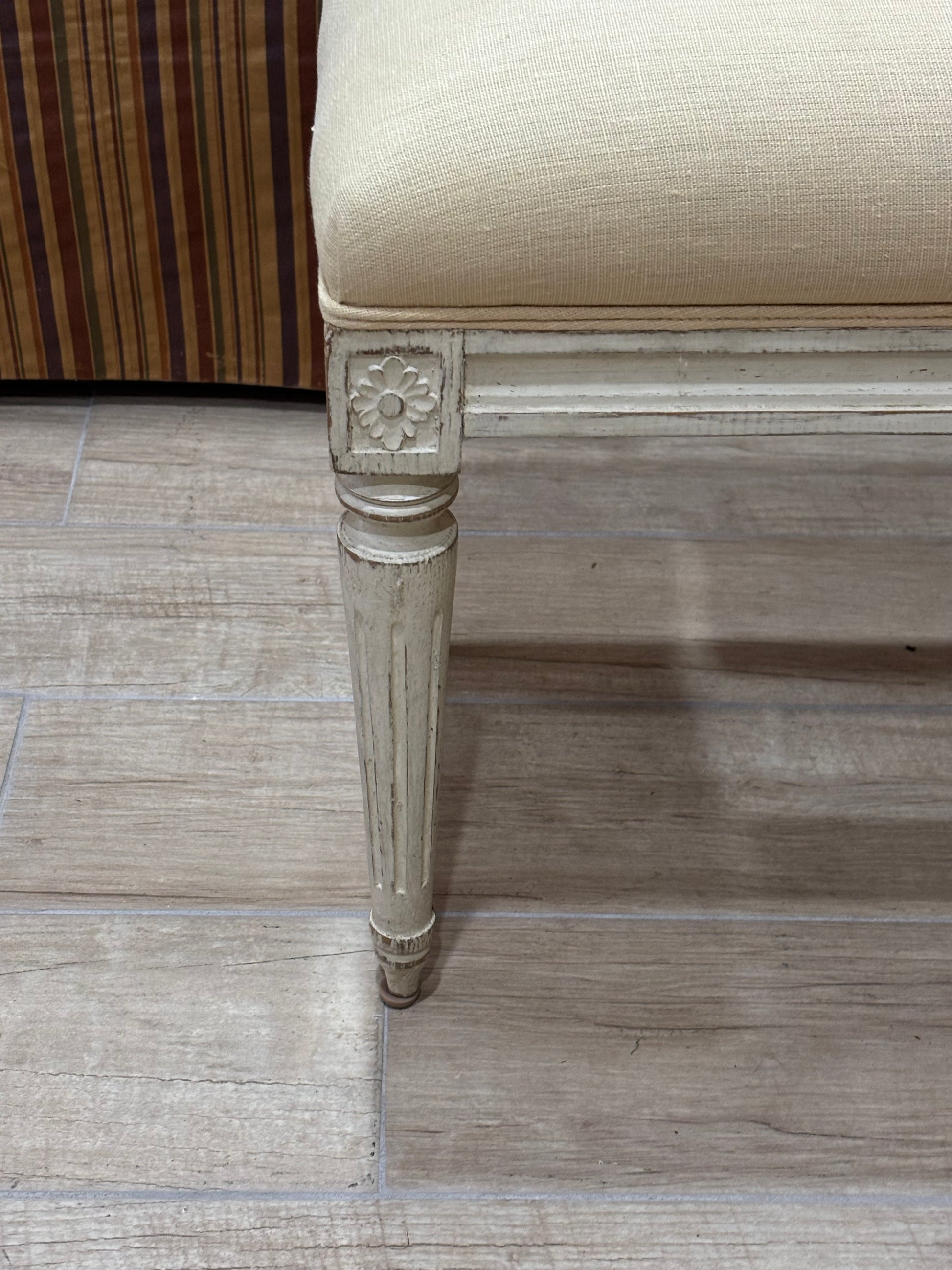 Close-up of a bench or stool leg with a distressed off-white painted frame and a beige upholstered seat, shown on a light wood-look floor.