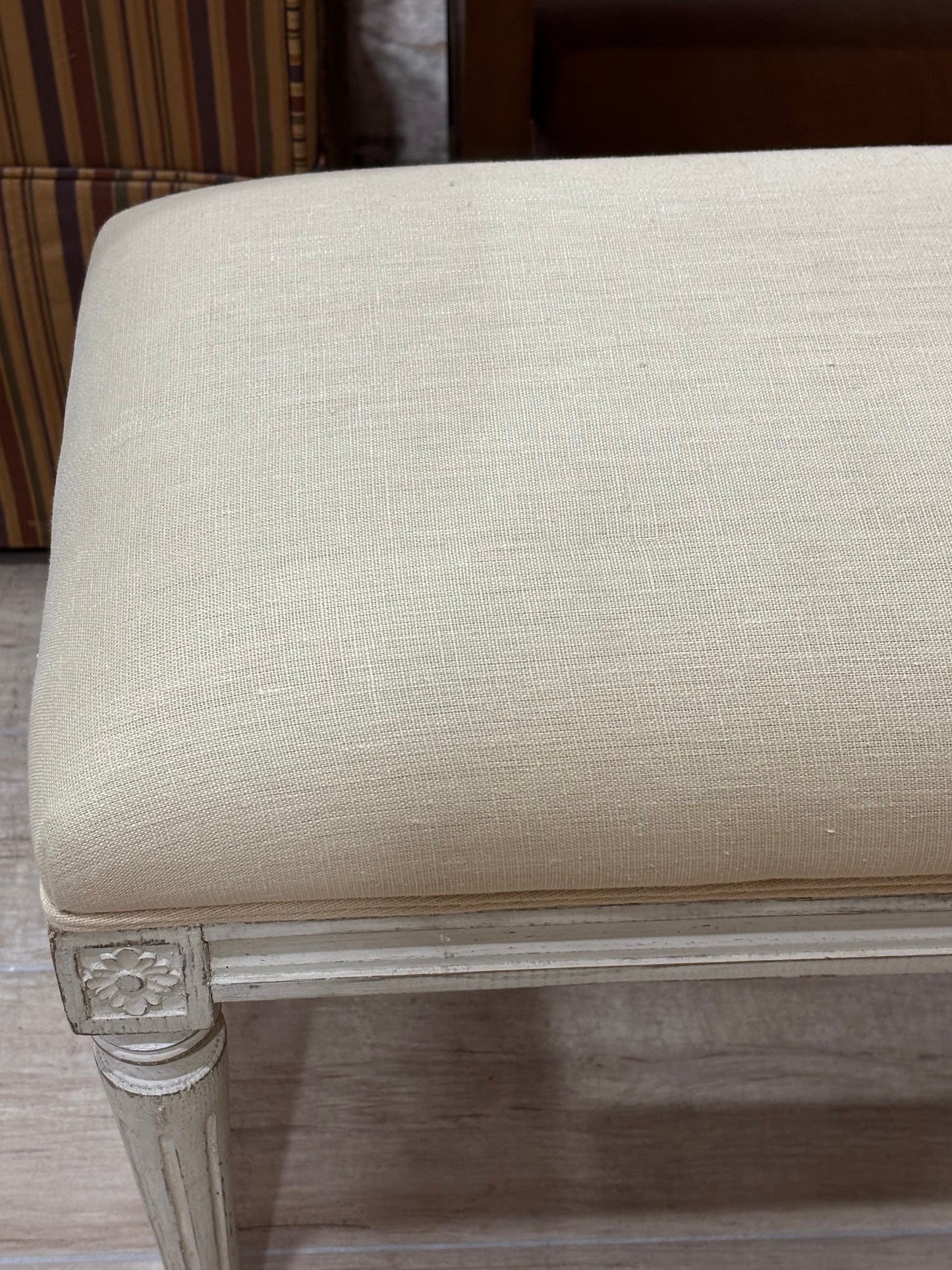 Close-up of a beige upholstered bench or ottoman with a textured woven fabric seat and a distressed off-white wooden frame with carved details on the leg.