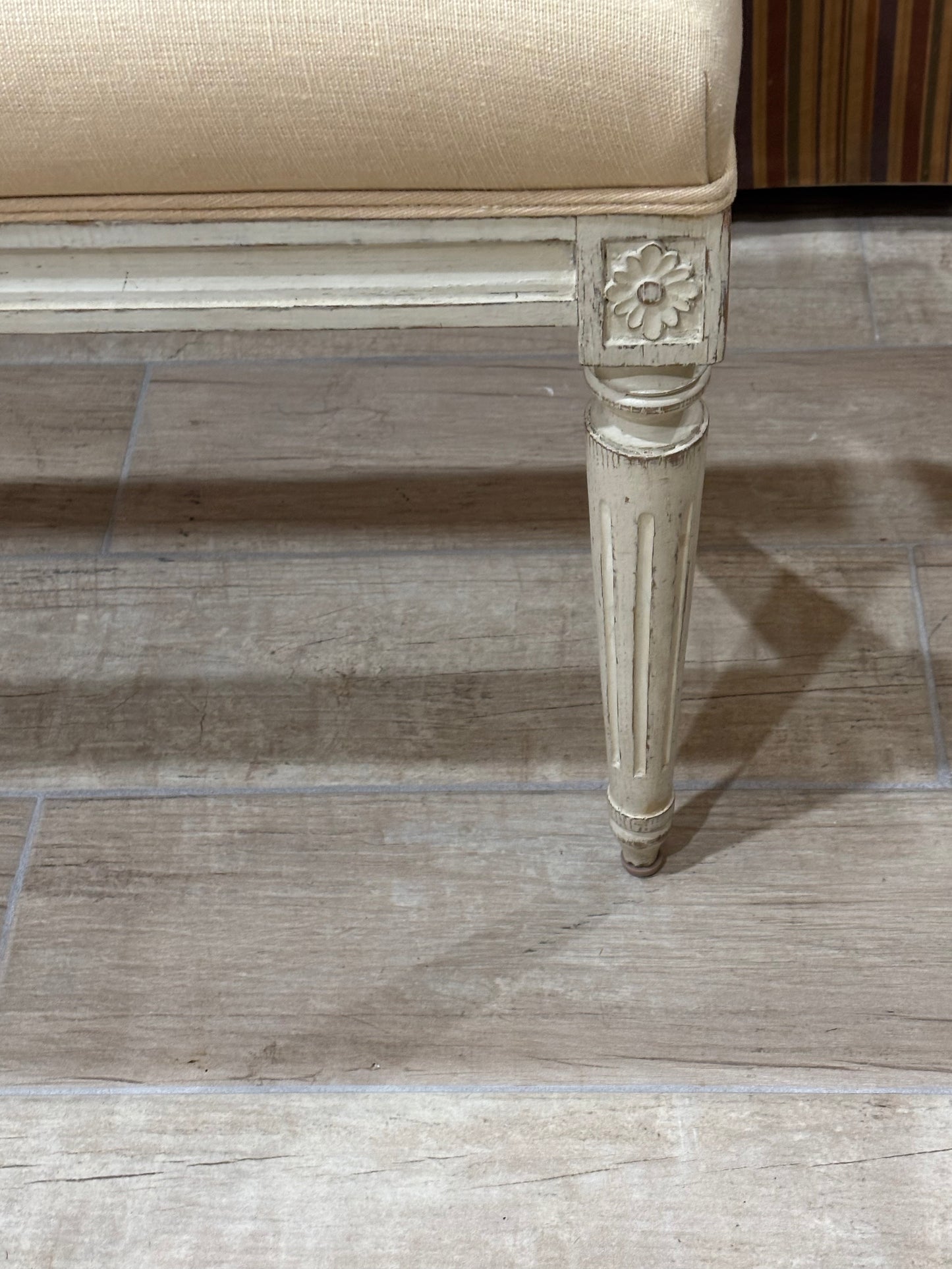 Close-up of a distressed cream upholstered bench or stool leg with a carved floral detail, set on a light wood-look tile floor.