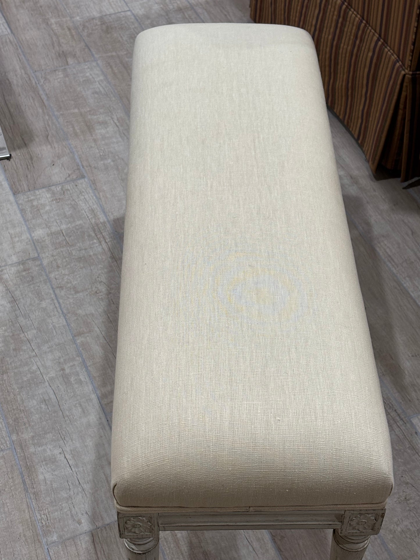 A long upholstered bench with a cream-beige textured fabric seat and carved wooden legs, shown on a gray wood-look floor.
