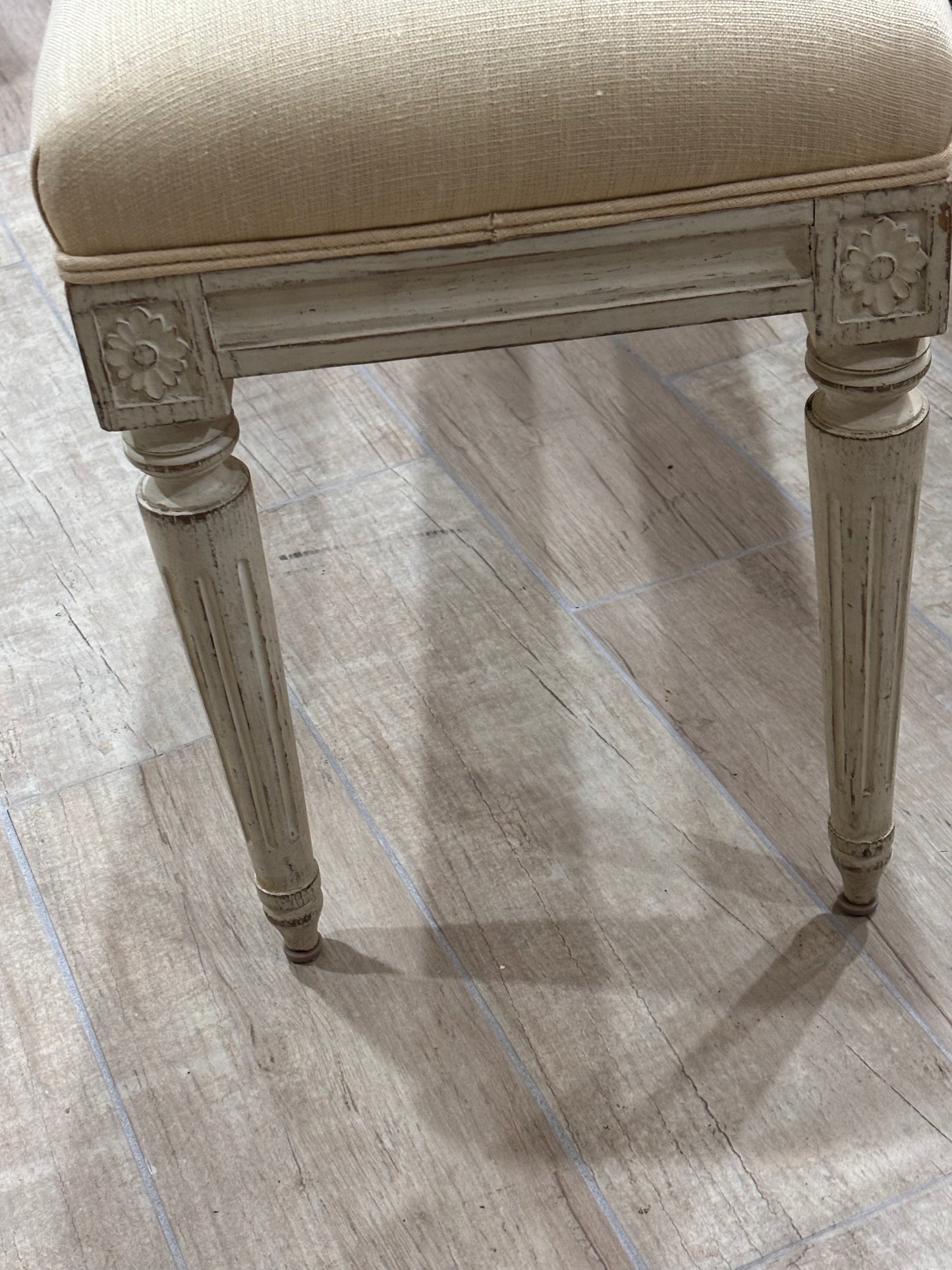 A close-up of a vintage-style upholstered stool with a distressed gray frame and a beige textured fabric seat, shown on a light wood-look tile floor.