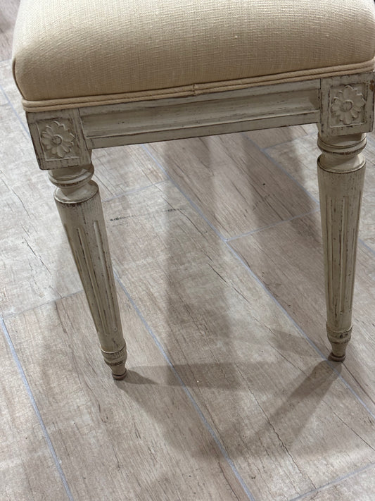 A close-up of a vintage-style upholstered stool with a distressed gray frame and a beige textured fabric seat, shown on a light wood-look tile floor.