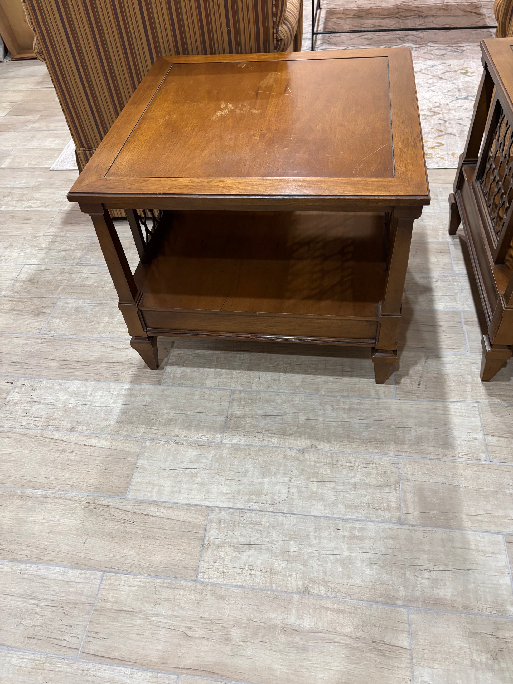 A square wooden end table with a lower shelf, made of medium brown wood, shown on a light beige tile floor next to other furniture.