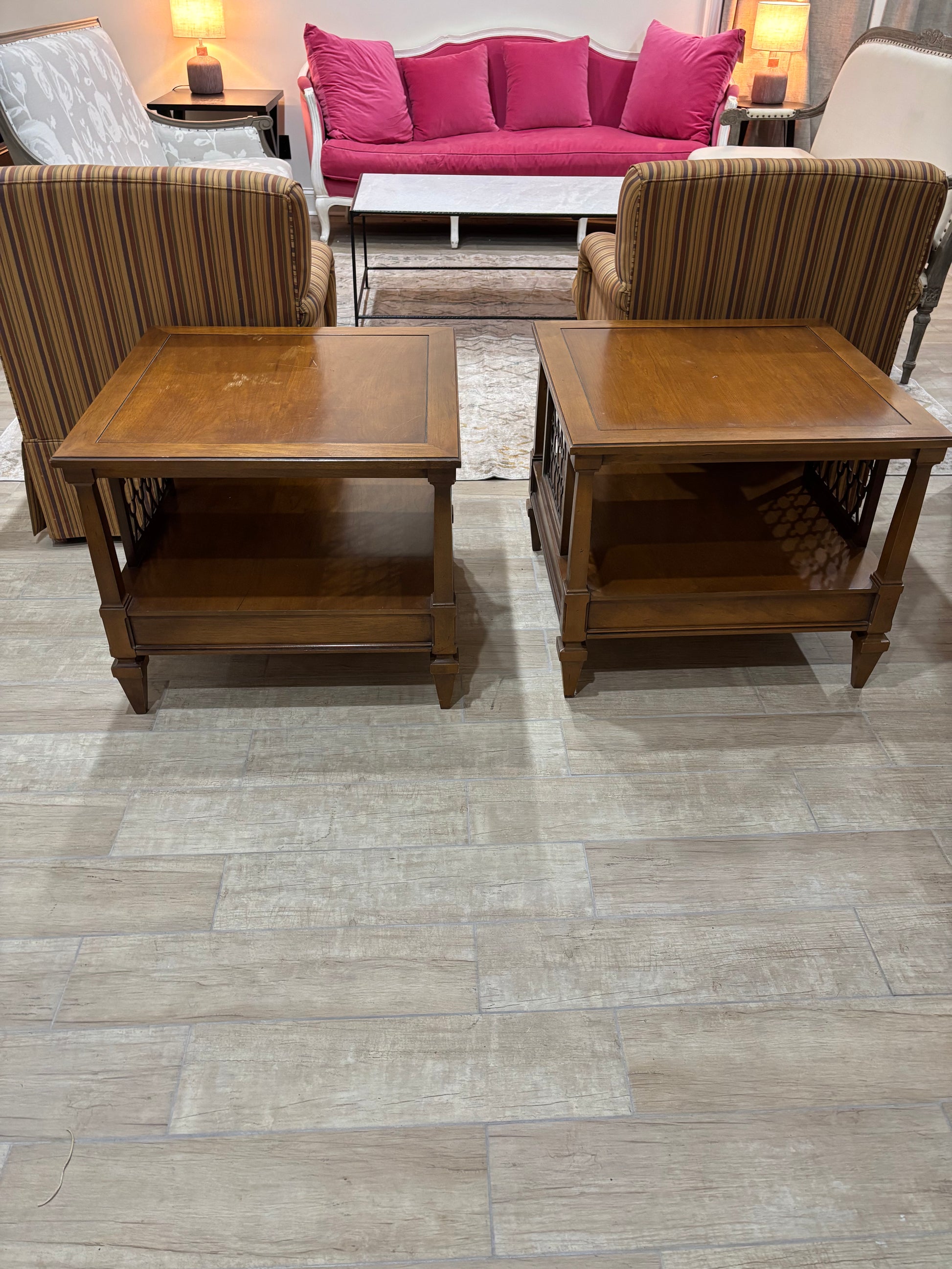 Two square wooden end tables with lower shelves, shown in a furniture showroom on a light gray wood-look floor. Striped armchairs and a bright pink sofa are visible in the background.