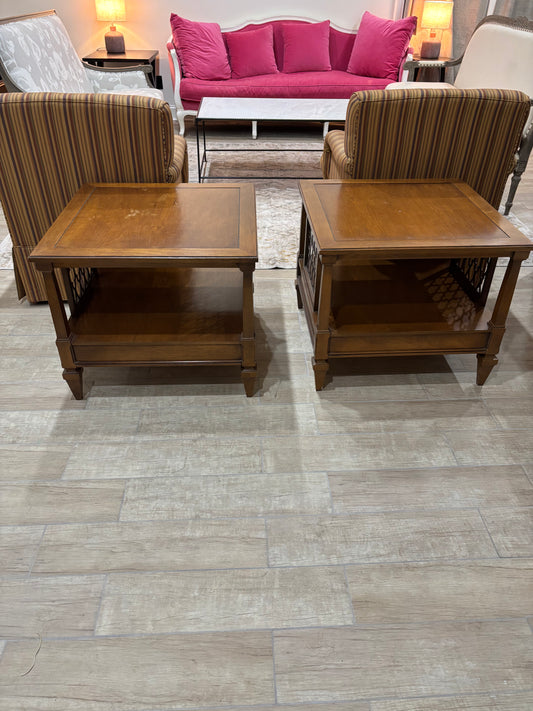 Two square wooden end tables with lower shelves, shown in a furniture showroom on a light gray wood-look floor. Striped armchairs and a bright pink sofa are visible in the background.