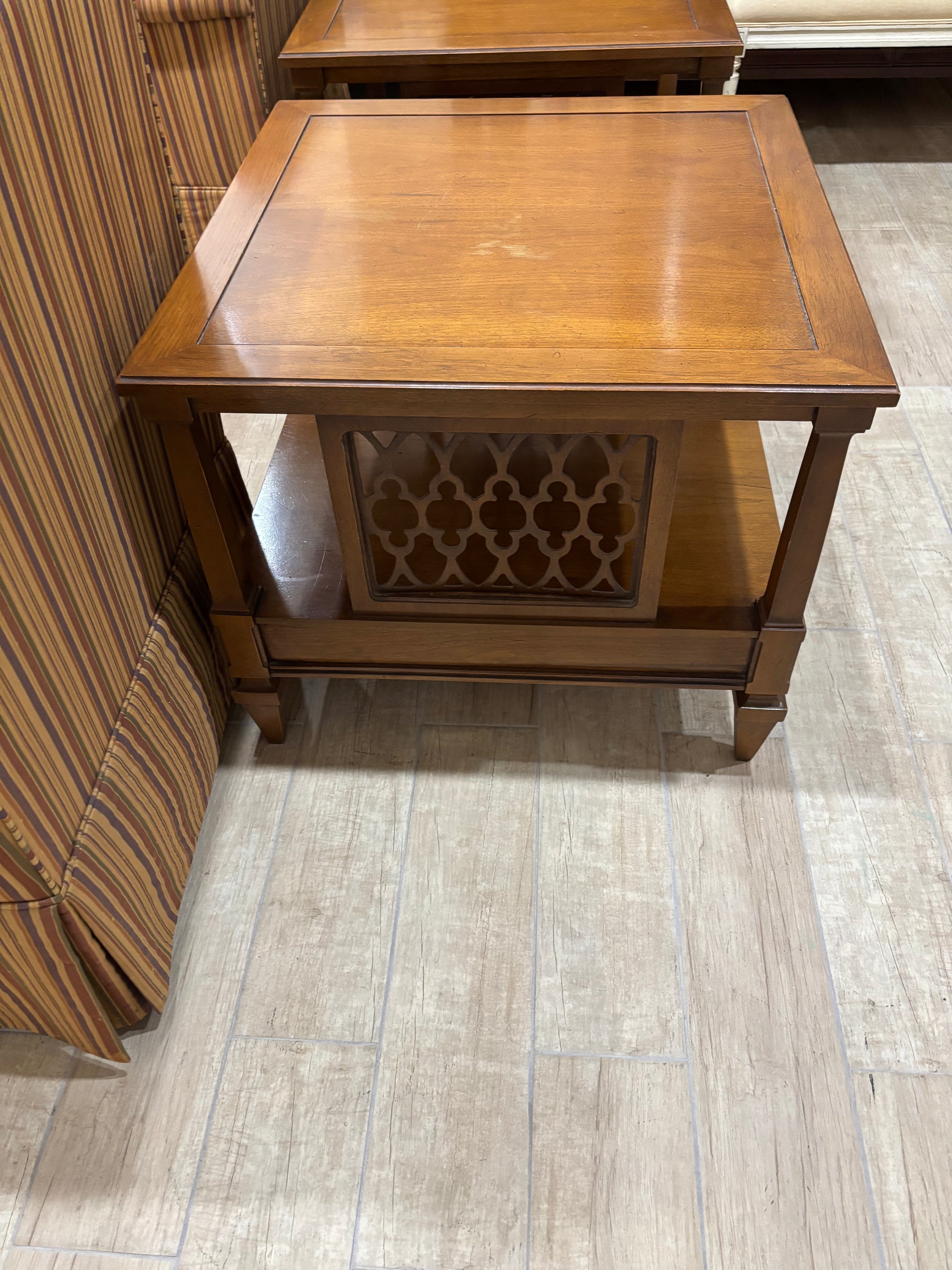 A square wooden end table with a warm medium-brown finish, featuring a decorative geometric lattice cutout panel on the front and a lower shelf, shown on a light wood-look floor next to a striped sofa.