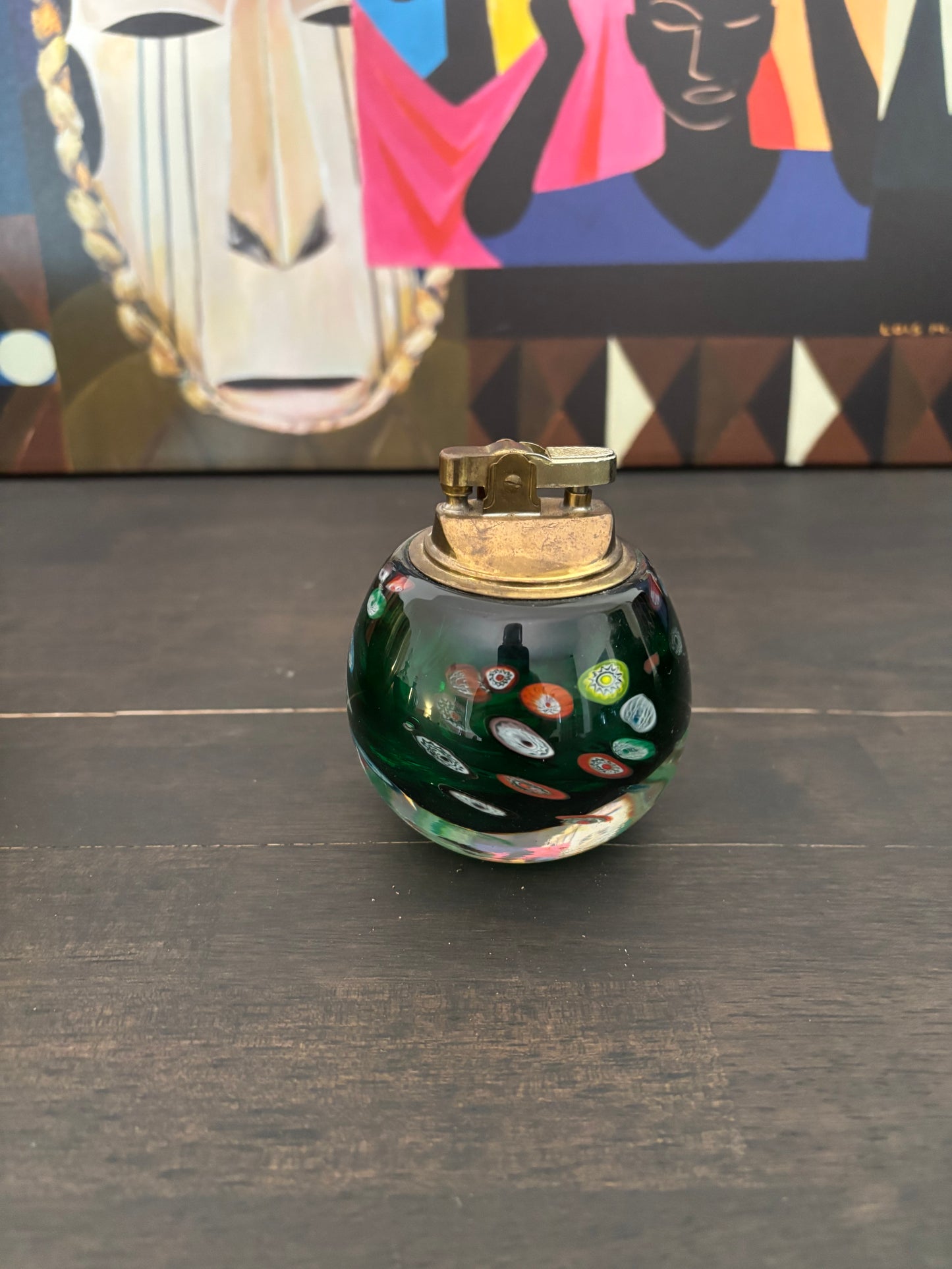 A small round glass bottle or perfume bottle with a gold-toned metal cap, featuring a dark green base with colorful glass inlay patterns. It is placed on a dark wooden surface with a colorful abstract art print in the background.