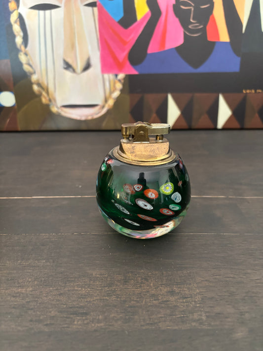 A small round glass bottle or perfume bottle with a gold-toned metal cap, featuring a dark green base with colorful glass inlay patterns. It is placed on a dark wooden surface with a colorful abstract art print in the background.