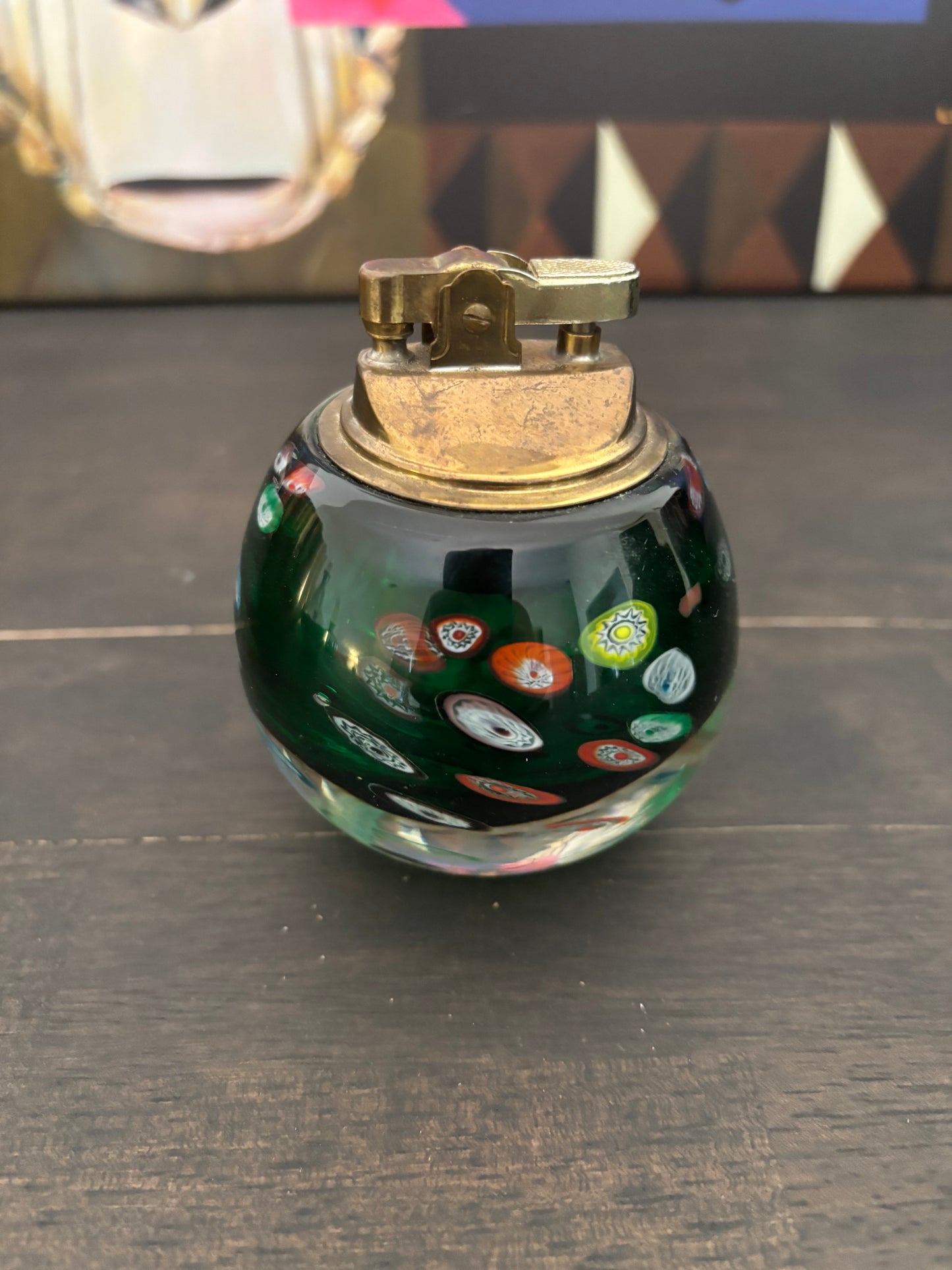 A small decorative glass lighter with a dark green body featuring colorful circular millo-fiori patterns, topped with a brass-colored metal ignition mechanism, resting on a dark wooden surface.