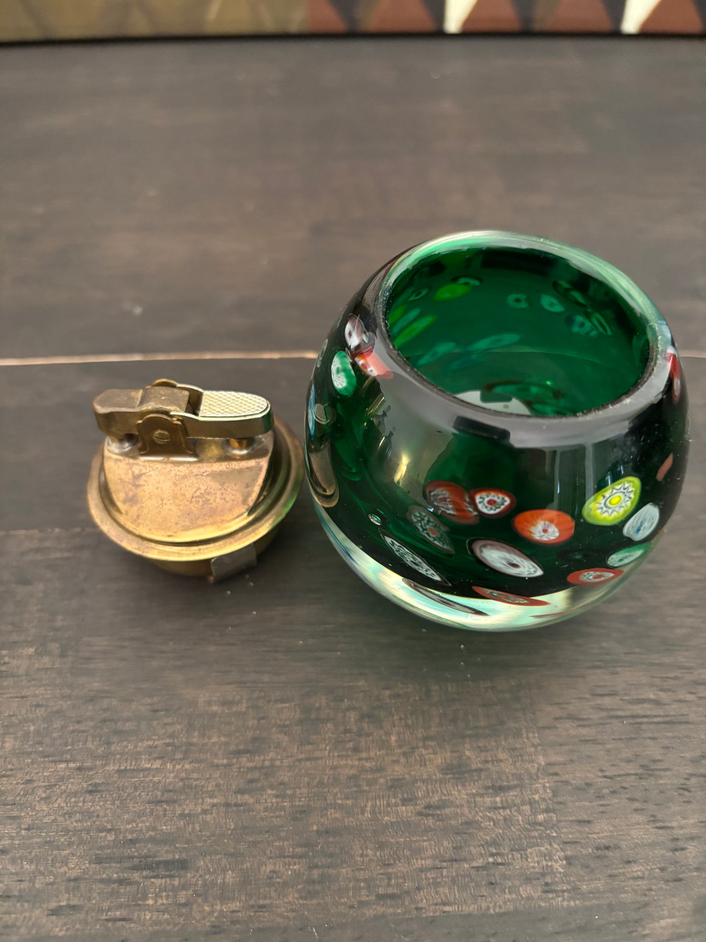 A small round green glass container with colorful bubble details and a separate brass lighter placed beside it on a dark wooden surface.