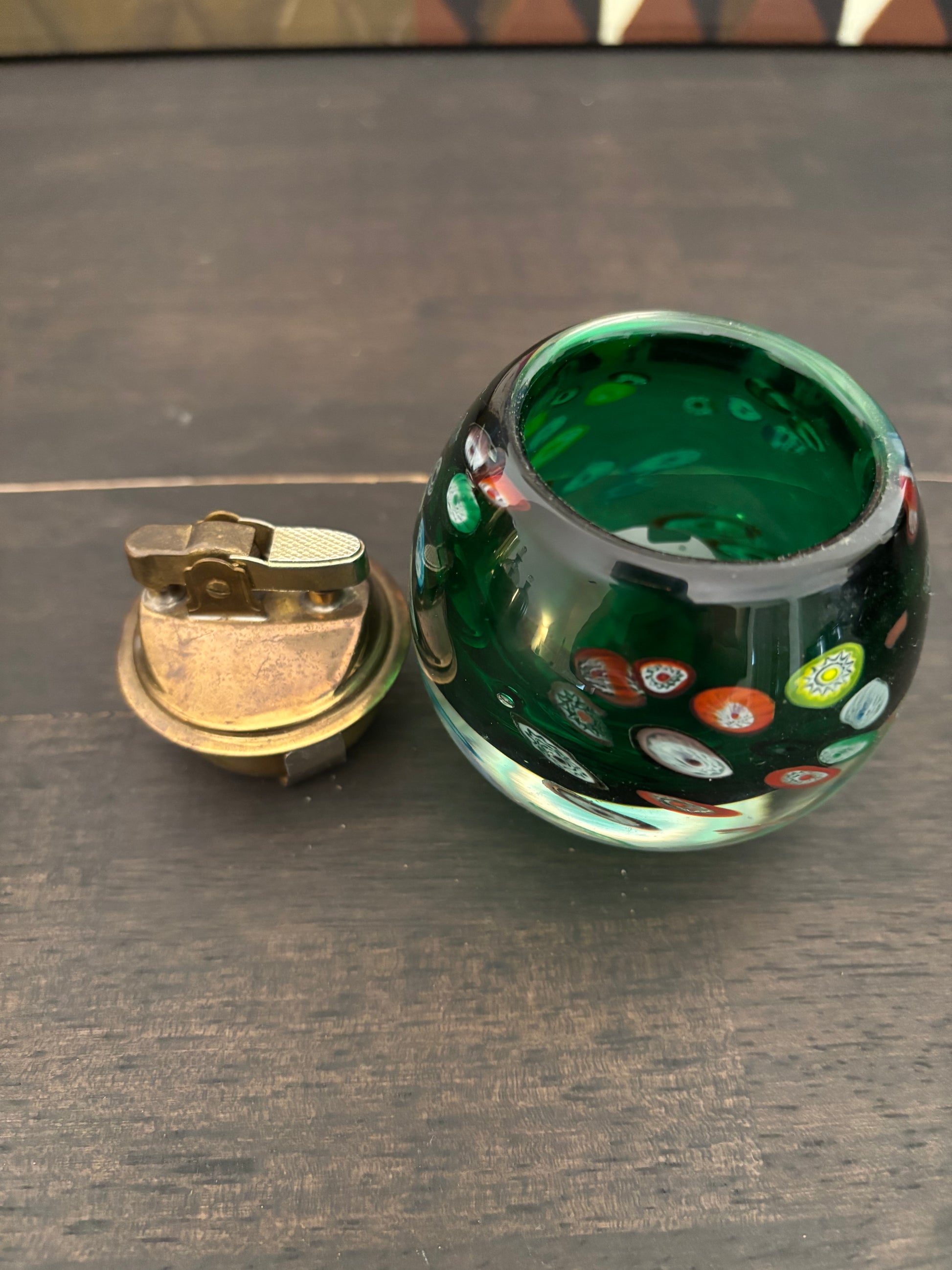 A small round green glass container with colorful bubble details and a separate brass lighter placed beside it on a dark wooden surface.