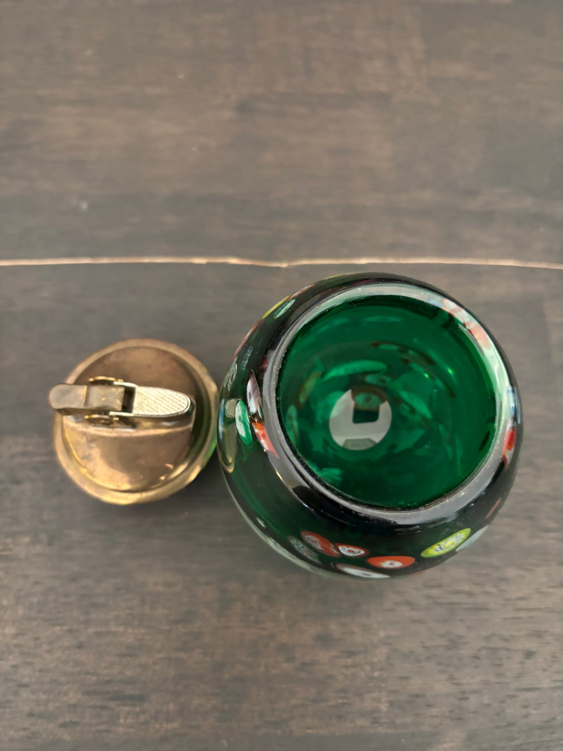 A close-up top-down view of a green glass container or candle holder with a separate brass-toned metal lid resting beside it on a dark wood surface.